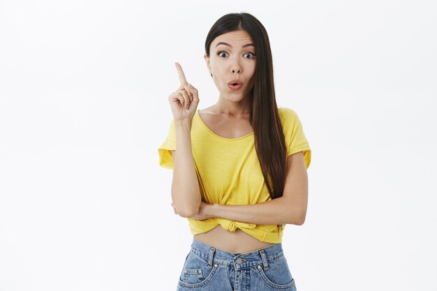 Espantada e animada, jovem atraente e elegante modelo feminina em uma camiseta amarela recortada com longos cabelos escuros dobrando os lábios levantando o dedo indicador