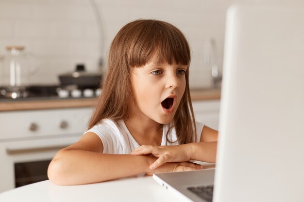 Espantada e espantada, criança do sexo feminino de cabelos escuros, sentada à mesa, olhando para a tela do laptop com a boca totalmente aberta, vê algo surpreendente, posando na cozinha.