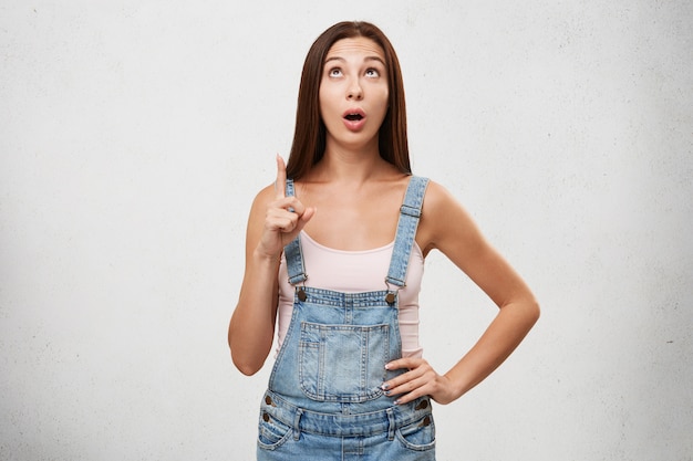 Espantado espantada mulher adolescente bonita vestindo macacão elegante olhando para cima em fascinante e apontando o dedo indicador, indicando algo curioso e chocante.