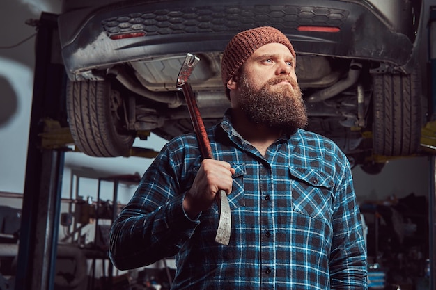 Especialista mecânico barbudo vestido com uma camisa de lã e gorro, de pé com um pé de cabra na mão, contra um carro em um elevador na garagem.