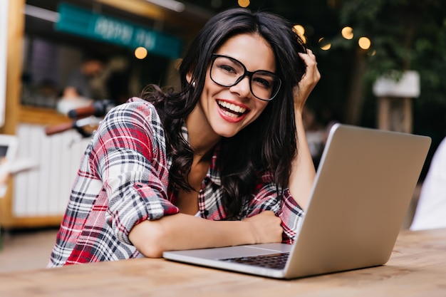 Espectacular jovem com cabelos escuros, sorrindo durante o trabalho com o computador. Foto ao ar livre de emocional aluna de óculos escuros.