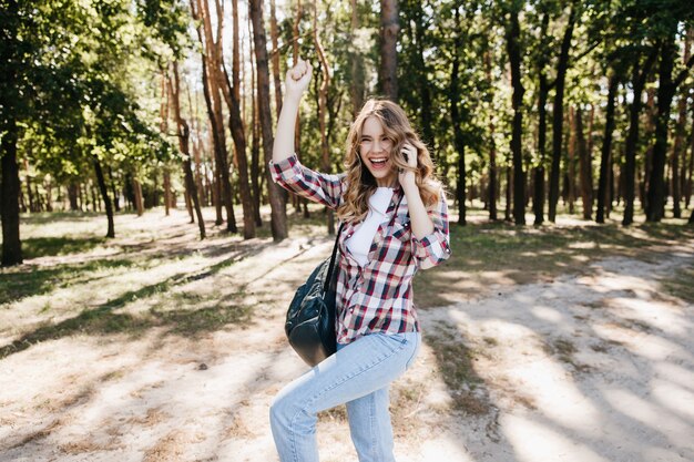 Espectacular jovem em jeans dançando na floresta de verão. Garota despreocupada com mochila brincando durante a sessão de fotos ao ar livre.