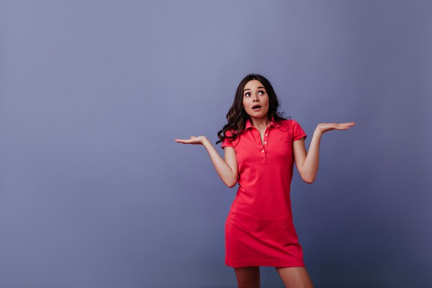 Espectacular jovem vestida de vermelho, posando com as mãos para cima. Garota de cabelos castanhos surpresa em pé na parede roxa com a boca aberta.