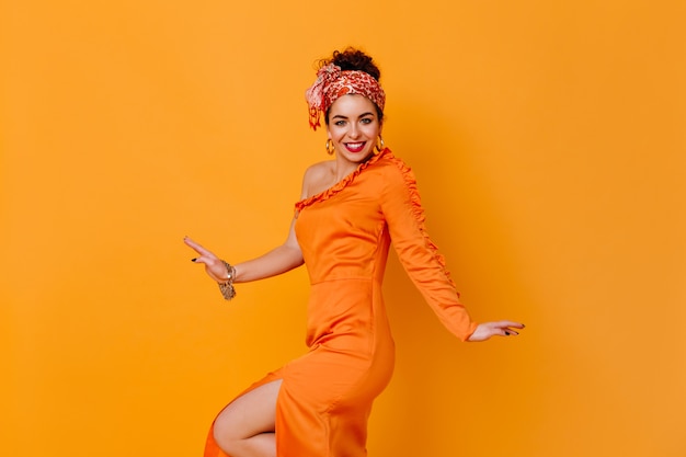 Foto grátis espectacular senhora com uma faixa incomum e vestido de cetim com fenda está sorrindo no espaço laranja.