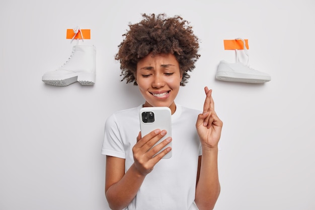 Foto grátis esperançoso cabelo encaracolado mantém o dedo cruzado morde os lábios aguarda por algo importante olha esperançosamente para a tela em poses de smartphone contra fundo branco com sapatos engessados. aguardando resultados