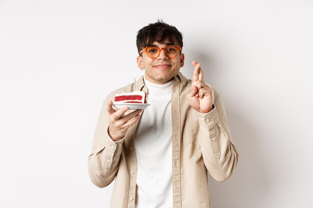 Esperançoso cara bonito de óculos, fazendo desejo no bolo de aniversário, em pé com os dedos cruzados e um sorriso feliz no fundo branco.