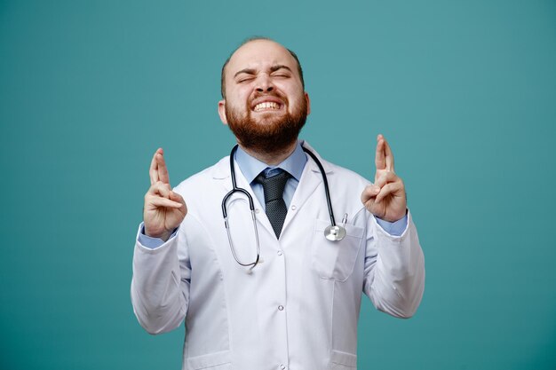 Esperançoso jovem médico masculino vestindo jaleco e estetoscópio em volta do pescoço fazendo desejo com os olhos fechados e dedos cruzados isolados no fundo azul