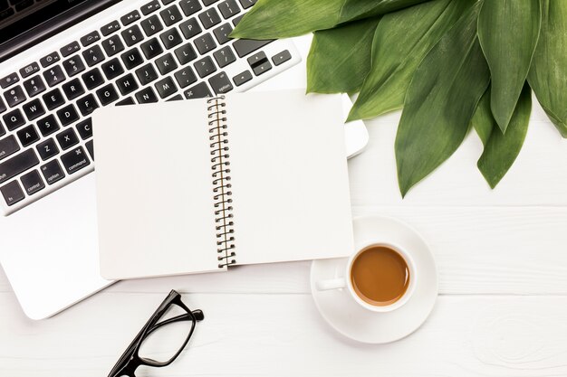 Espiral notepad e folhas no laptop com óculos e xícara de café na mesa de escritório de madeira