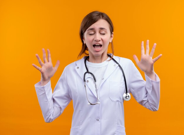 Espirrando, jovem médica vestindo bata médica e estetoscópio, mostrando as mãos vazias com os olhos fechados na parede laranja