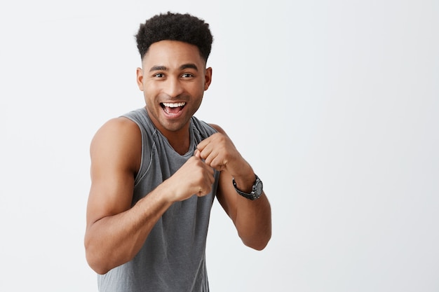 Esporte é divertido. Retrato de alegre preto bonito homem de pele com penteado afro na camisa cinza desportiva, sorrindo brilhantemente, posando para o photoshoot de jornal de universidade, mostrando o esporte é saudável e divertido