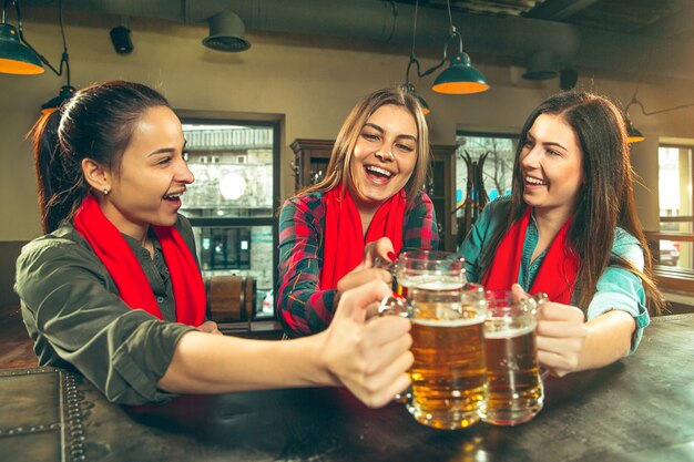 Esporte, pessoas, lazer, amizade, conceito de entretenimento - fãs de futebol feminino felizes ou bons jovens amigos bebendo cerveja, comemorando a vitória em um bar ou pub. Conceito de emoções positivas humanas