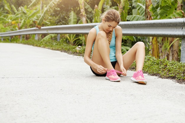 Foto grátis esportes e conceito de estilo de vida saudável. jovem desportiva sentada na estrada amarrando o tênis rosa durante o exercício de corrida ao ar livre.