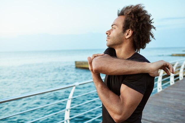 Esportes, fitness e estilo de vida saudável. Atleta afro-americano em boa forma, parece concentrado enquanto estica os braços à beira-mar, fazendo exercícios de alongamento de braços e ombros