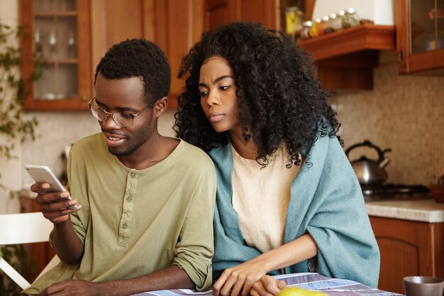 Esposa negra suspeita tentando ler a mensagem que seu feliz marido envia para alguém no celular, enquanto ela suspeita de traição, não confiando nele. Ciúme, infidelidade e desconfiança