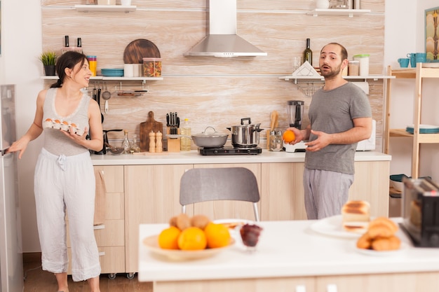 Esposa tirando ovos da geladeira para fazer café da manhã para ela e o marido na cozinha. Marido conversando com a esposa enquanto ela prepara ovos para o café da manhã.