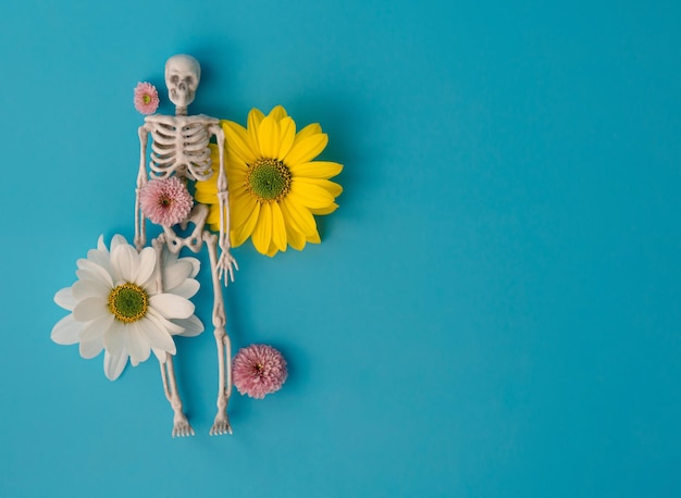 Esqueleto de brinquedo em flores de crisântemo brancas e amarelas sobre fundo azul, criativo conceito halloween, a ideia de vida e morte, espaço livre para texto
