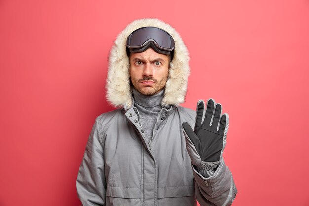 Esquiador descontente sério com roupas de inverno usa óculos de esqui na cabeça mantém a palma da mão para frente faz o gesto de pare.