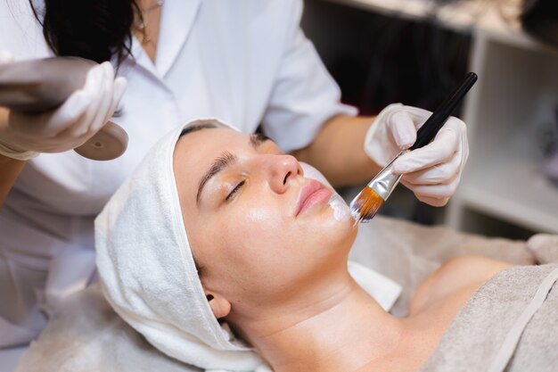 Esteticista com um pincel aplica uma máscara hidratante branca no rosto de uma jovem cliente em um salão de beleza de spa