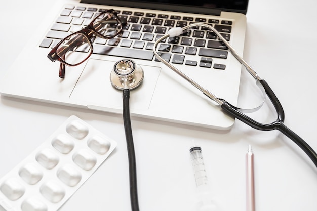 Estetoscópio e óculos no teclado do laptop com pacote de medicina; seringa e caneta em fundo branco