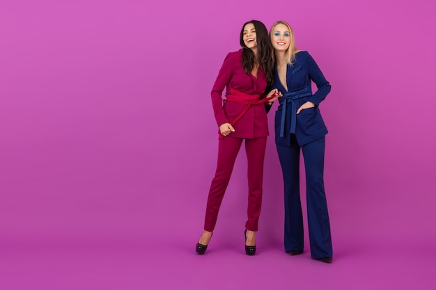 Foto grátis estilo de alta costura duas mulheres atraentes sorridentes na parede violeta em elegantes ternos coloridos de cor roxa e azul, amigos se divertindo juntos, tendência da moda