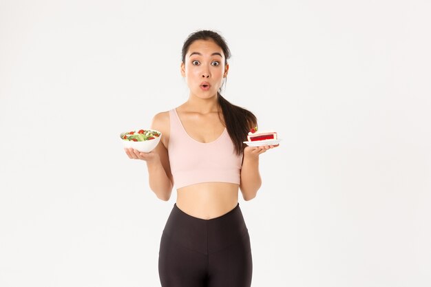 Estilo de vida ativo, conceito de fitness e bem-estar. Retrato de indecisa e tentadora linda garota asiática tentando resistir à tentação de segurar um bolo delicioso, estar de dieta, olhando para uma salada saudável.