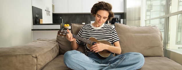 Foto grátis estilo de vida e hobbies conceito jovem mulher sorridente no sofá tocando ukulele cantando e aprendendo