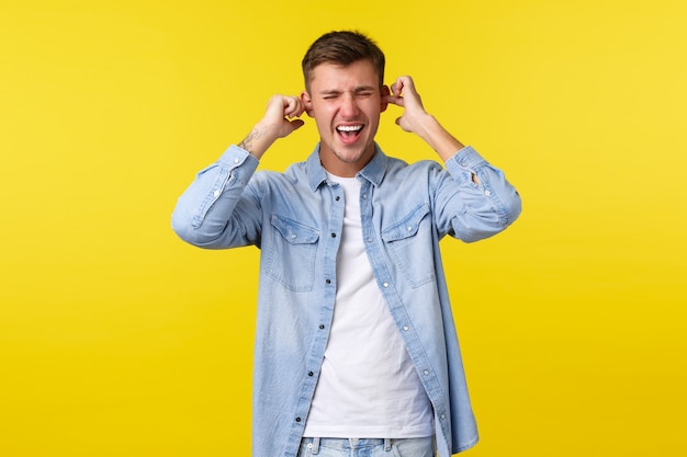 Estilo de vida, emoções das pessoas e conceito de lazer de verão. Aluno bonito angustiado e irritado gritando irritado, orelhas fechadas com os dedos, sem vontade de ouvir, fundo amarelo de pé.