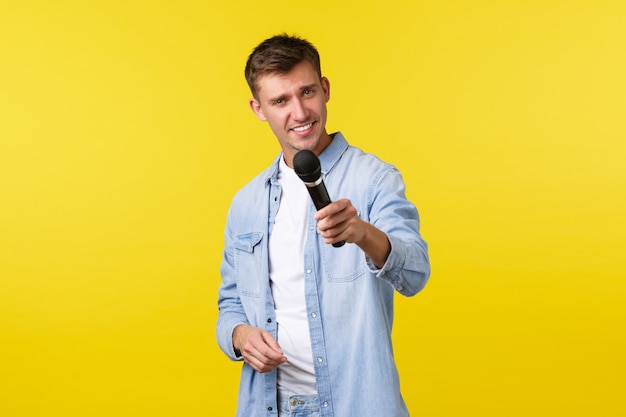 Estilo de vida, emoções das pessoas e conceito de lazer de verão. Louro atrevido e bonito performer entregando o microfone, entrevistando alguém, sorrindo atrevidamente e mostrando fundo amarelo