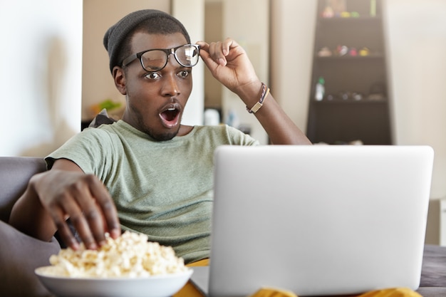 Estilo de vida moderno, tecnologia e conceito de pessoas. Homem afro-americano jovem atônito relaxando em casa depois do trabalho assistindo jogo de basquete on-line ou vídeos nas mídias sociais e comendo pipoca