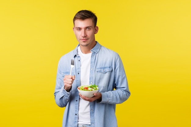 Foto grátis estilo de vida saudável, pessoas e conceito de comida. louro atrevido comendo salada, apertando os olhos e sorrindo encantado, cozinhando um café da manhã vegan, em pé com um fundo amarelo satisfeito