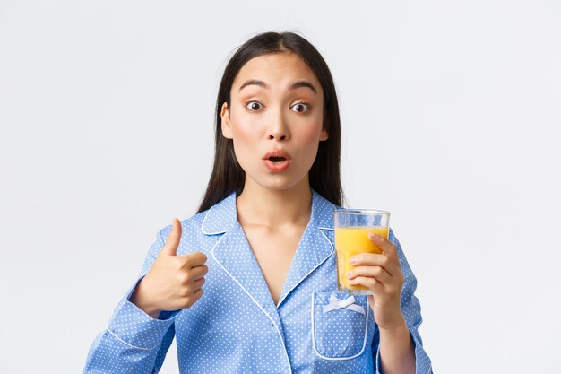 Estilo de vida saudável, rotina matinal e conceito de pessoas. Close-up de menina asiática surpresa e satisfeita parecendo espantado como mostrando o polegar para cima e tentando suco de laranja fresco, fundo branco.