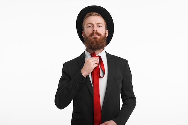 Estilo, moda masculina e conceito de moda. Foto de um homem caucasiano elegante com uma espessa barba ruiva se vestindo para um evento oficial, usando um chapéu preto e terno, amarrando uma elegante gravata vermelha