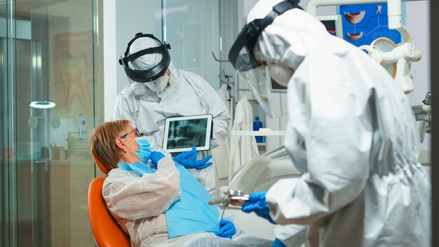 Estomatologista em traje de proteção revisando radiografia de dente com paciente sênior, explicando o tratamento com comprimido na pandemia de covisd-19. Equipe médica usando protetor facial, macacão, máscara e luvas.