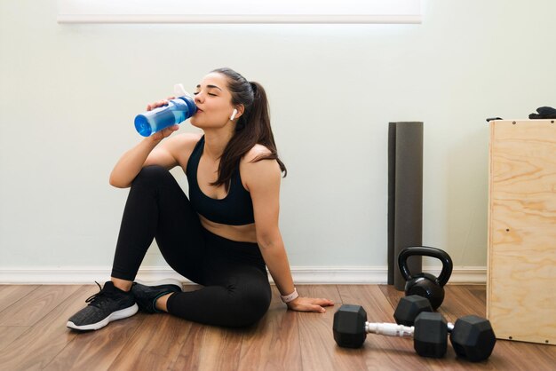 Estou com tanta sede. Mulher jovem e bonita com fones de ouvido ouvindo música enquanto bebe água depois de suar durante o treino cardio em casa