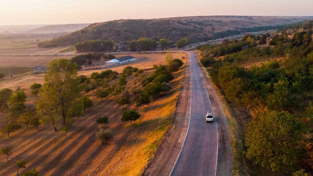 Estrada rural e carro em movimento ao nascer do sol, campos, colinas cobertas de árvores