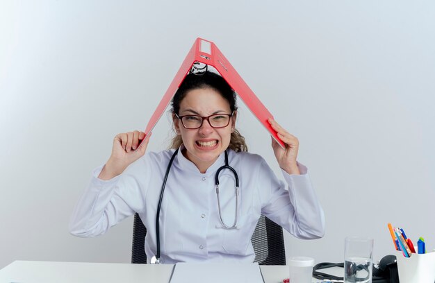 Estressada jovem médica vestindo túnica médica, estetoscópio e óculos, sentada na mesa com ferramentas médicas, olhando segurando uma pasta na cabeça isolada