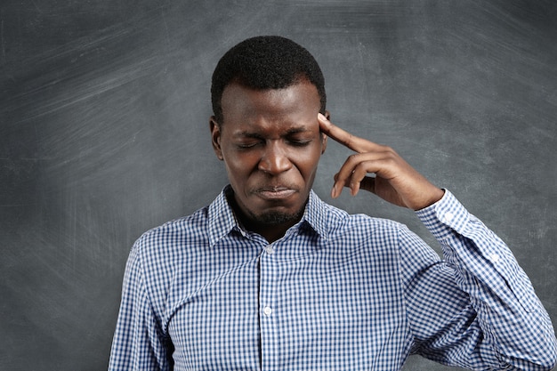 Foto grátis estressado empresário africano com expressão meticulosa lutando para se lembrar de algo, fechando os olhos com força e pressionando o dedo na têmpora como se estivesse com forte dor de cabeça.