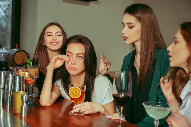 Estresse. Amigos do sexo feminino tomando uma bebida no bar. Eles estão sentados em uma mesa de madeira com coquetéis. Eles estão vestindo roupas casuais. Amigos confortando e acalmando uma garota chorando