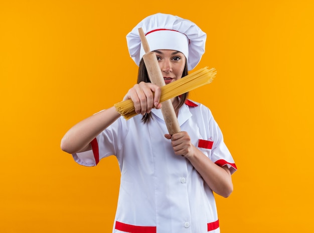 estrita jovem cozinheira vestindo uniforme de chef segurando e cruzando espaguete com rolo de massa isolado na parede laranja