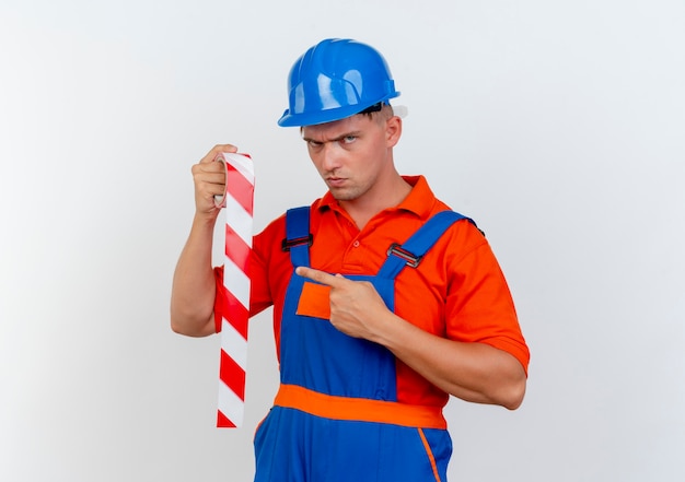 Estrito jovem construtor do sexo masculino usando uniforme e segurando capacete de segurança e aponta para fita adesiva em branco