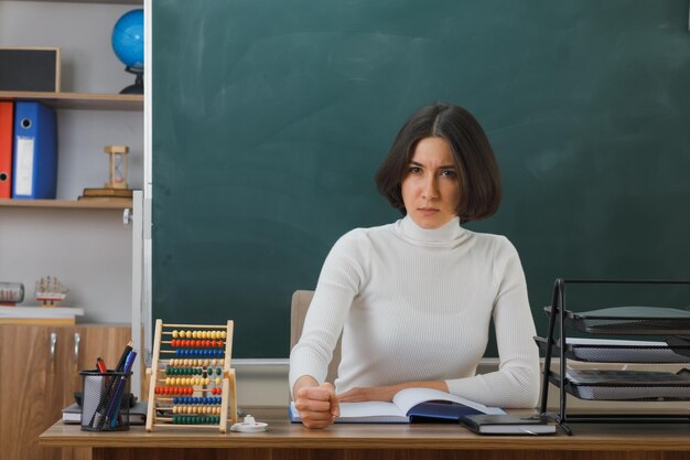 estrito olhando para a câmera jovem professora sentada na mesa com ferramentas escolares na sala de aula