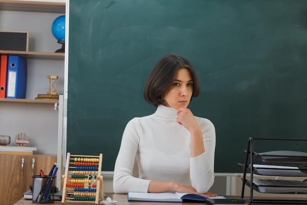 Estrito queixo agarrado jovem professora sentada na mesa com ferramentas escolares na sala de aula
