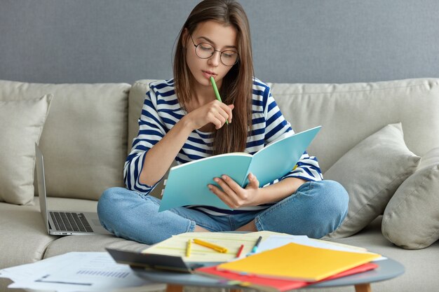 Estudando o conceito online. Mulher jovem séria ocupada com um projeto freelance remoto, senta-se em um sofá confortável, faz anotações, segura o livro, usa o laptop em casa com internet sem fio