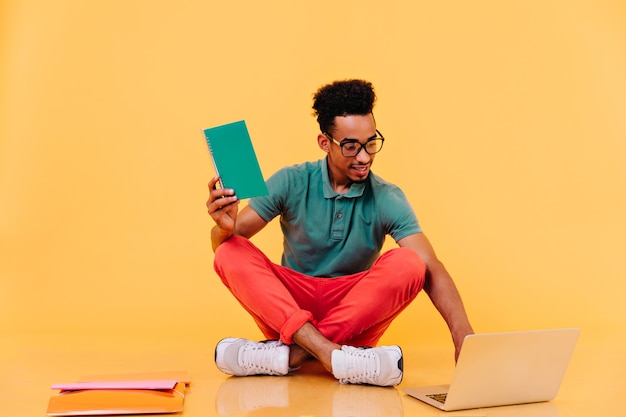Estudante africano interessado em copos, sentado no chão com livros didáticos. freelancer masculino em roupas brilhantes, usando o laptop.