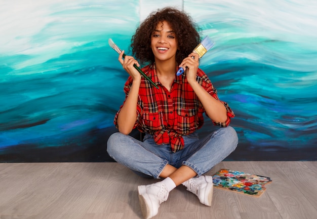 Estudante bonito bonito etnia africana segurando escovas e posando sobre suas obras de arte abstratas na lona.