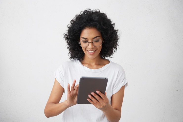 Foto grátis estudante carismática e bonita sorridente de pele escura, segurando um gadget moderno, usando o tablet para videochamada com os amigos, assistindo vídeos engraçados ou fazendo lição de casa, conversando