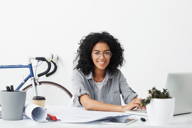 Estudante de design sorridente, sentada em seu local de trabalho, cercada de dispositivos