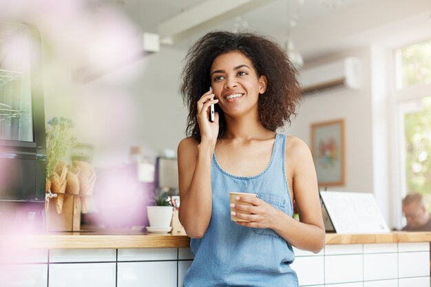 Estudante de mulher africana jovem alegre bonita sorrindo falando no telefone, bebendo café no café.