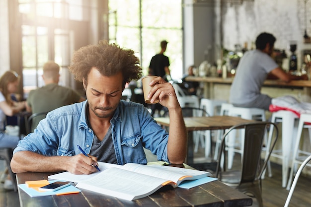 Estudante de pele escura do sexo masculino com penteado africano encaracolado, fazendo as tarefas de casa, preparando-se para a redação da aula no caderno, tomando café no refeitório, parecendo sério, concentrado