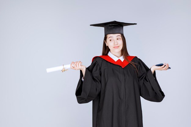 Estudante de pós-graduação em vestido segurando o celular e o diploma. Foto de alta qualidade